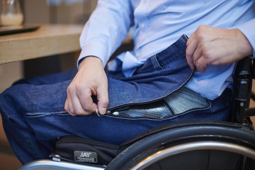 A male wheel chair user zipping his adaptive designed jeans. The zip is positioned on the side of the trouser 