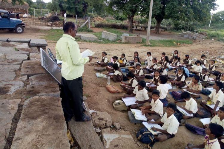 a male teacher in a village setting standing in front of a group of students who are sitting on the floor, during a class session